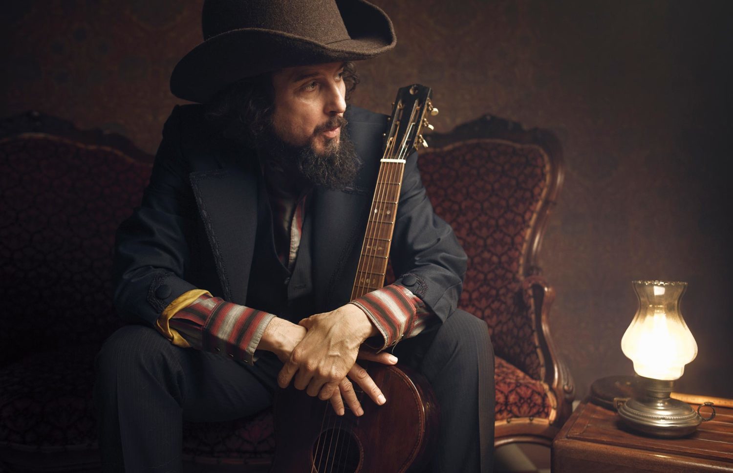 Vinicio Capossela seduto su una poltrona con la chitarra fra le braccia e luce morbida da una lampada