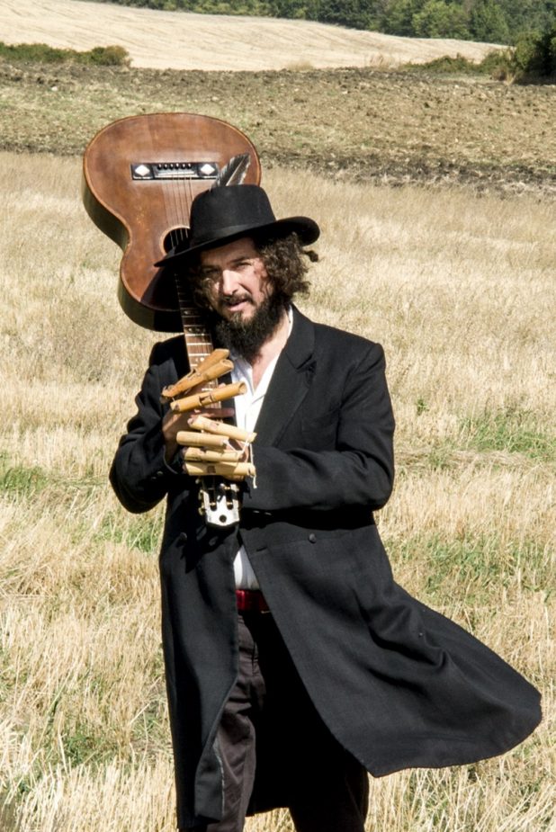 Vinicio Capossela ritratto con la chitarra mentre cammina su un campo di grano